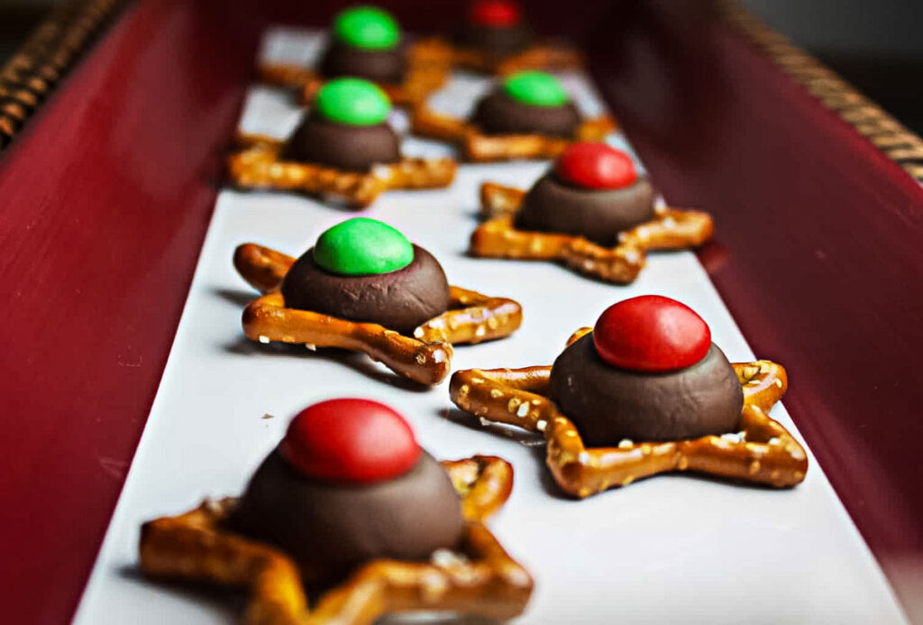 A row of star-shaped pretzels topped with chocolate kisses and red or green M&Ms on a rectangular white tray.