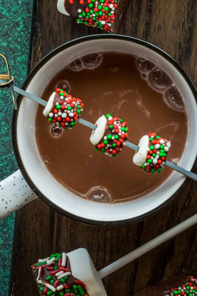 No-Bake Marshmallow Pops – Quick, Fun & Kid-Friendly a closeup of Mini Marshmallow pops with red and green sprinkles skewered and set on a cup of hot cocoa