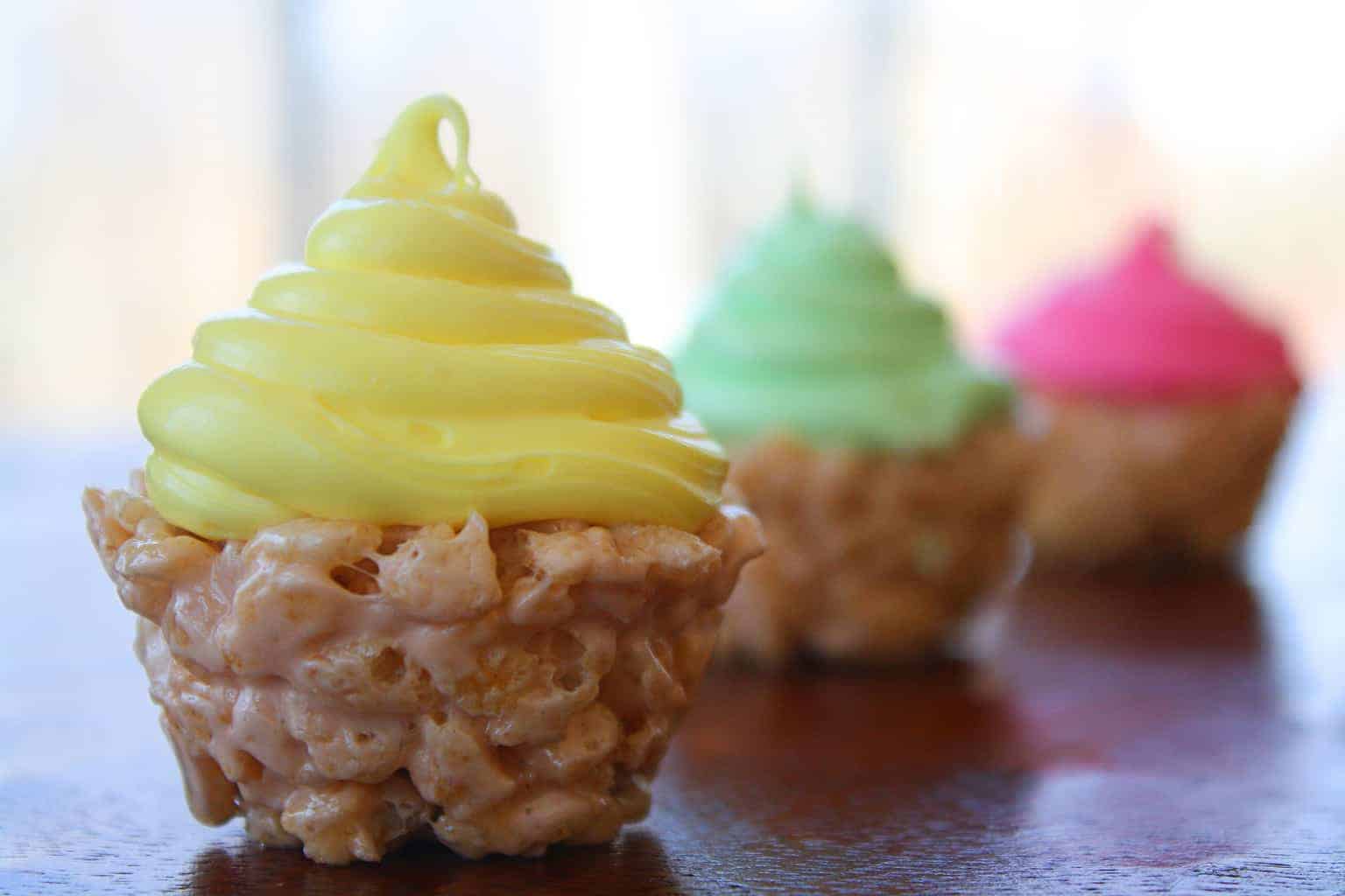 rice krispies cupcakes trio with yellow, green and pink frostings on a brown table next to a window