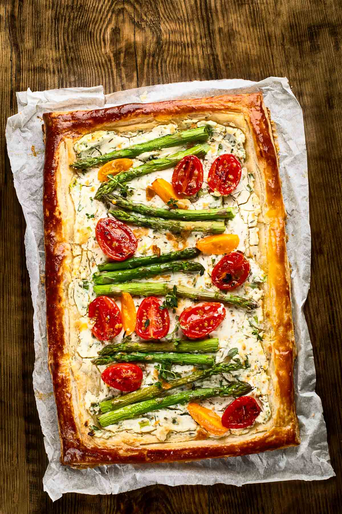 an asparagus goat cheese tart on parchment paper with halved cherry tomatoes and fresh herbs in puff pastry on a dark wood table