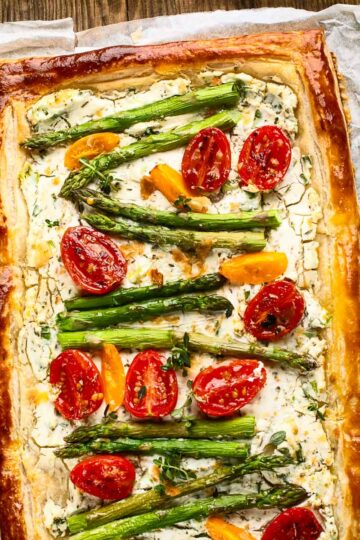 an asparagus goat cheese tart on parchment paper with halved cherry tomatoes and fresh herbs in puff pastry