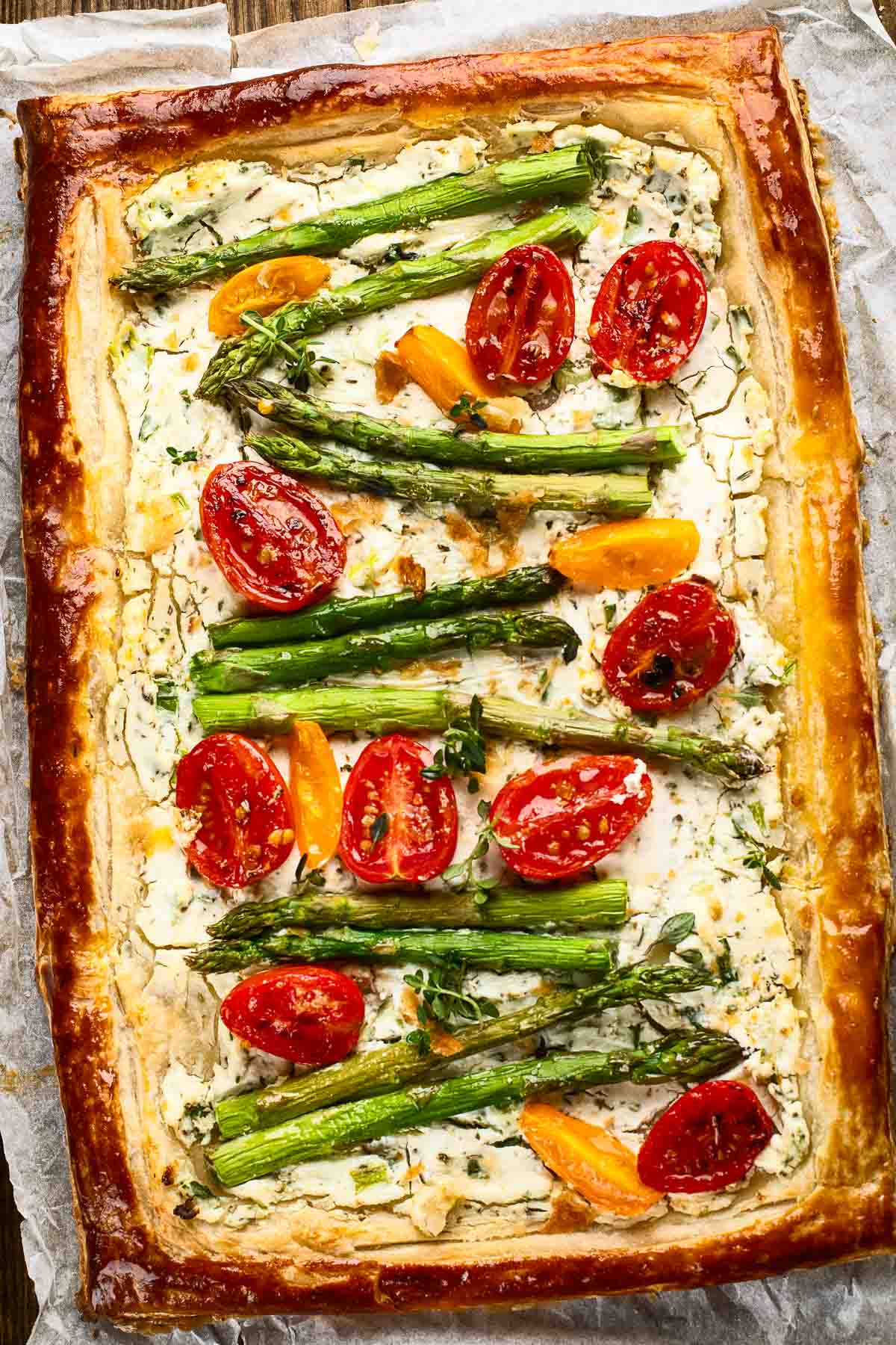 Asparagus Goat Cheese Tart an asparagus goat cheese tart on parchment paper with halved cherry tomatoes and fresh herbs in puff pastry