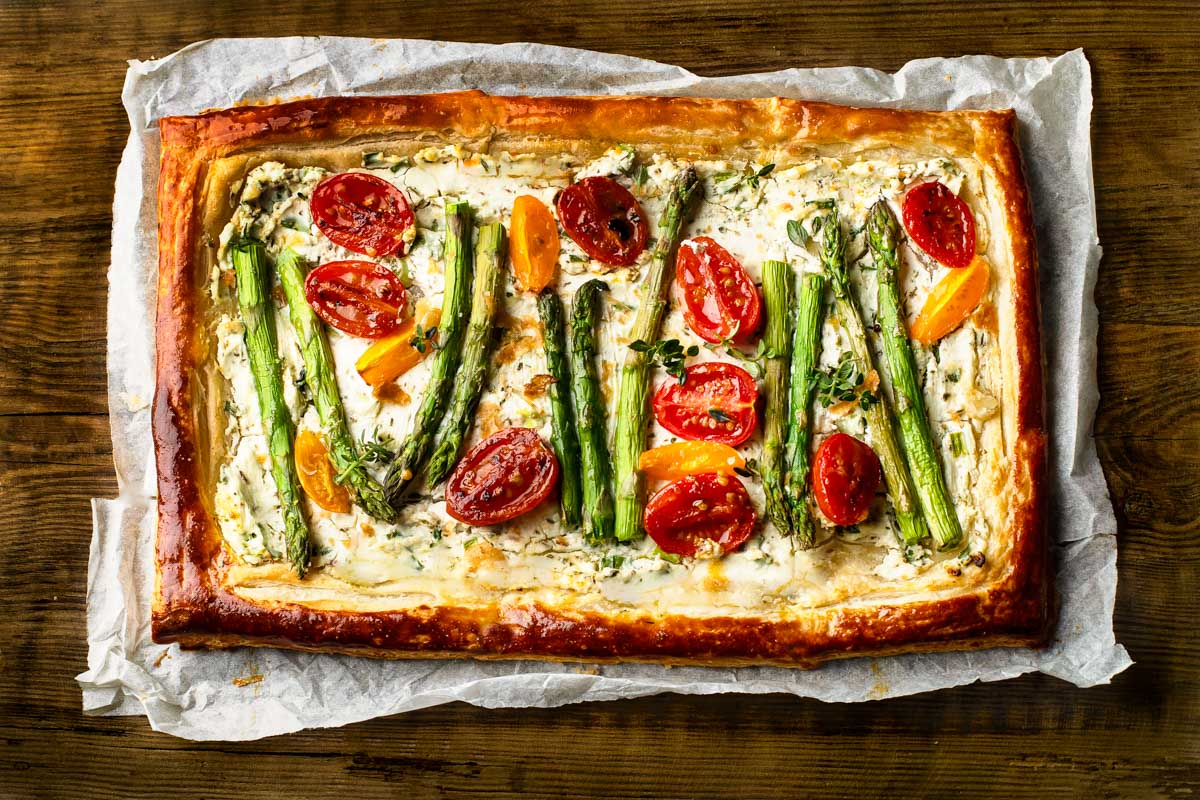 Asparagus Goat Cheese Tart side view of an asparagus goat cheese tart on parchment paper with halved cherry tomatoes and fresh herbs in puff pastry