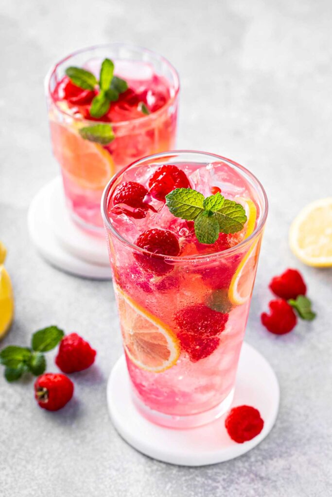 Homemade Raspberry Lemonade (Sweet, Tart & Refreshing) a glass of raspberry lemonade with thin lemon slices, fresh raspberries, and a sprig of fresh mint on white coasters with a few pieces of each fruit on the table