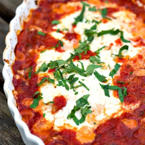 a white scalloped dish filled with goat cheese, marinara and fresh basil that's been baked