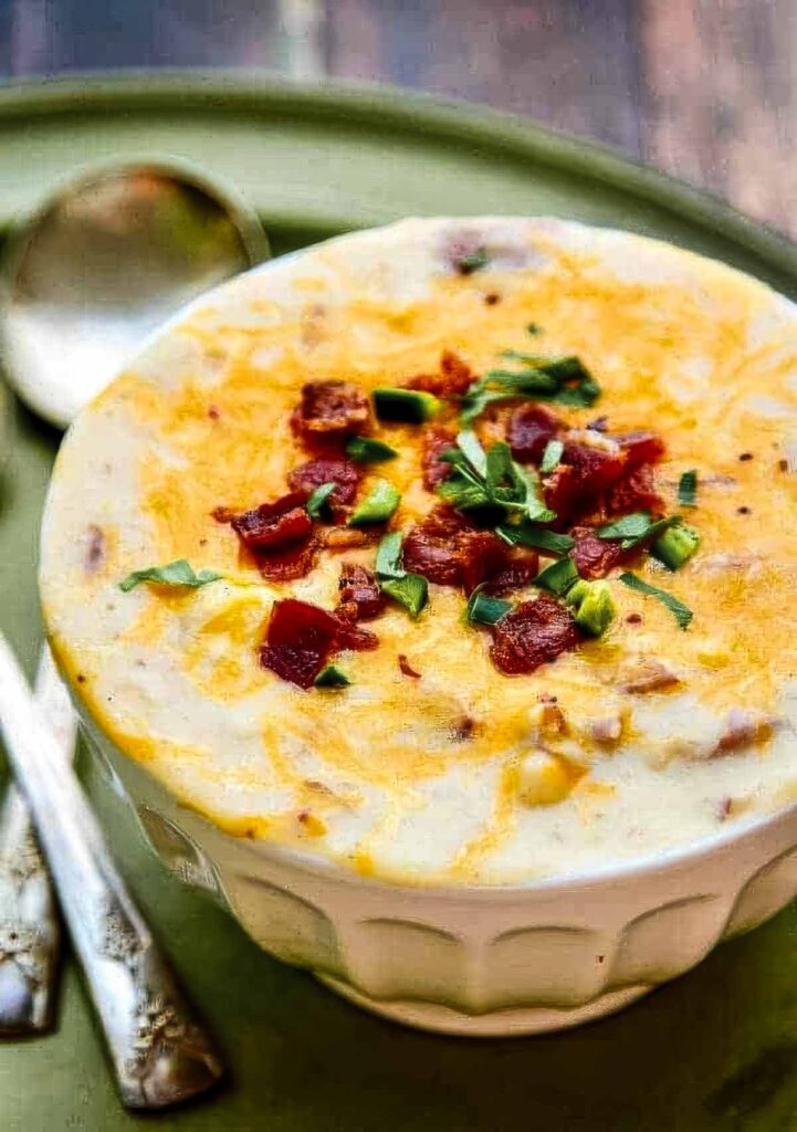 Bowl of thick and creamy potato soup topped with melted cheese, crispy bacon pieces, and fresh herbs on a rustic green plate.