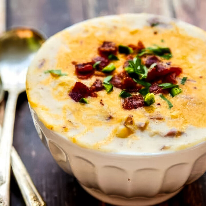 Bowl of loaded potato soup on a wooden table, topped generously with cheddar cheese, bacon bits, and chopped green onions, with spoons nearby.