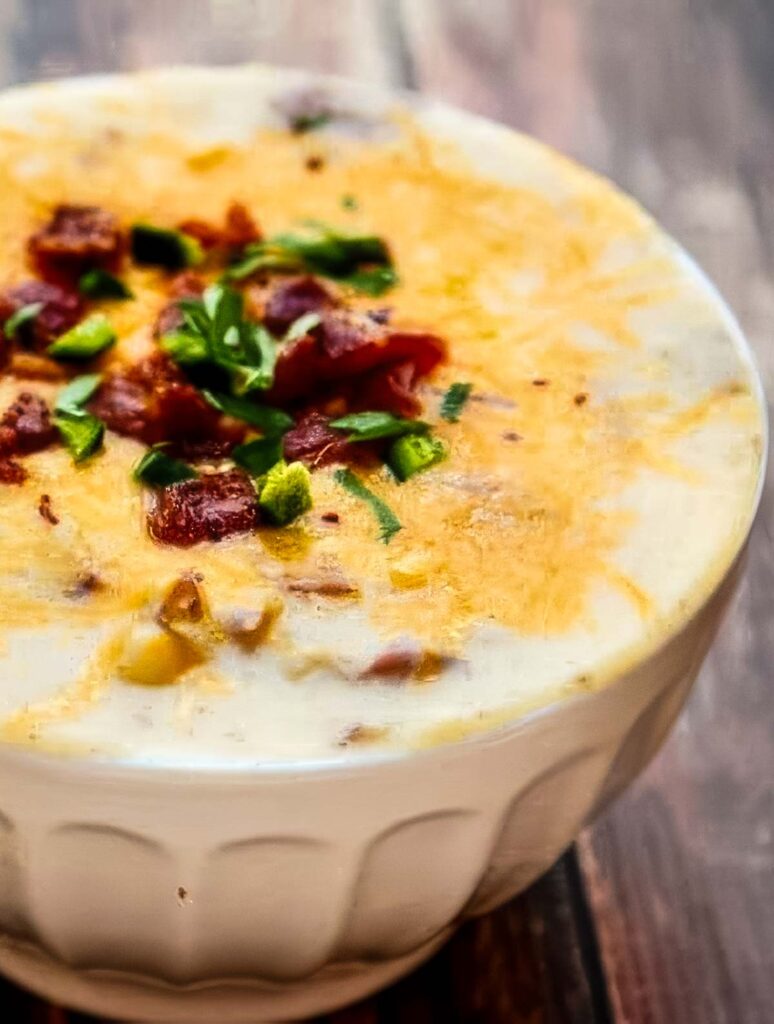 Side view of a bowl of loaded potato soup with visible potato chunks, melted cheese, bacon, and jalapeños, set on a rustic table.