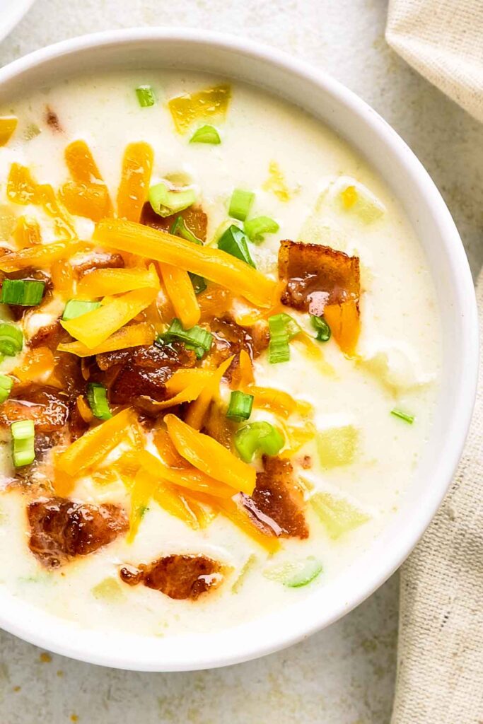 Bowl of loaded potato soup filled with chunks of potato, melted cheese, green onions, and bacon crumbles on a light background.