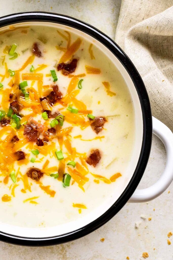 Close-up of creamy loaded potato soup in a white bowl with crispy bacon, shredded cheddar cheese, and sliced green onions on top.