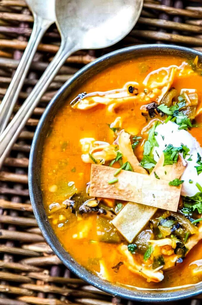 a bowl of chicken tortilla soup with hatch chiles that's topped with crispy tortilla strips, sour cream, and chopped fresh cilantro next to two spoons