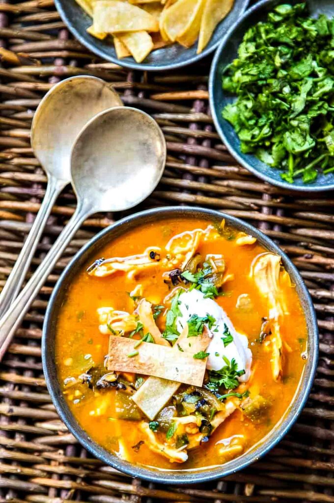 a bowl of chicken tortilla soup with hatch chiles that's topped with crispy tortilla strips, sour cream, and chopped fresh cilantro next to two spoons plus a small dish of cilantro and another of crispy tortilla strips