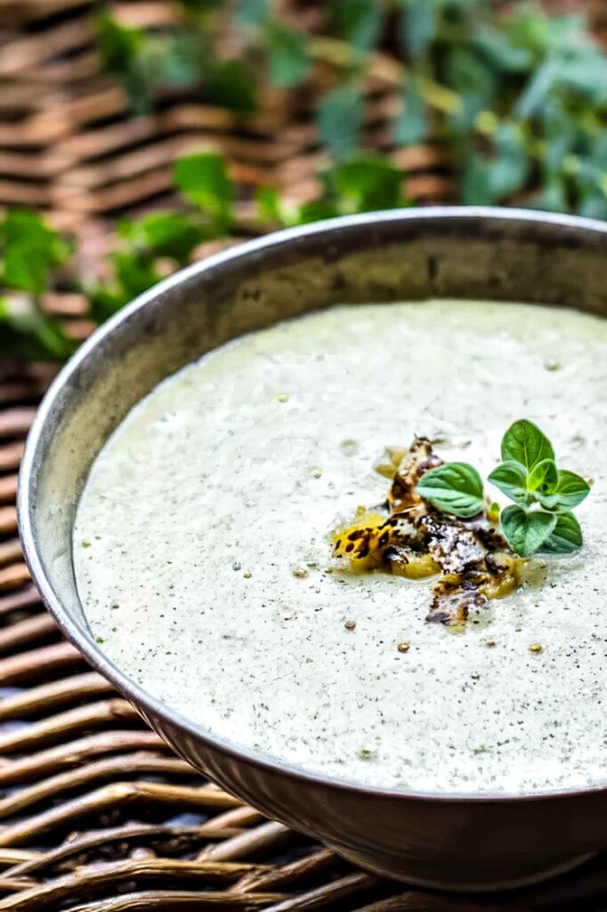 Creamy green Hatch Chile Enchilada Sauce in a bowl on a woven mat, topped with charred chiles and a sprig of oregano.