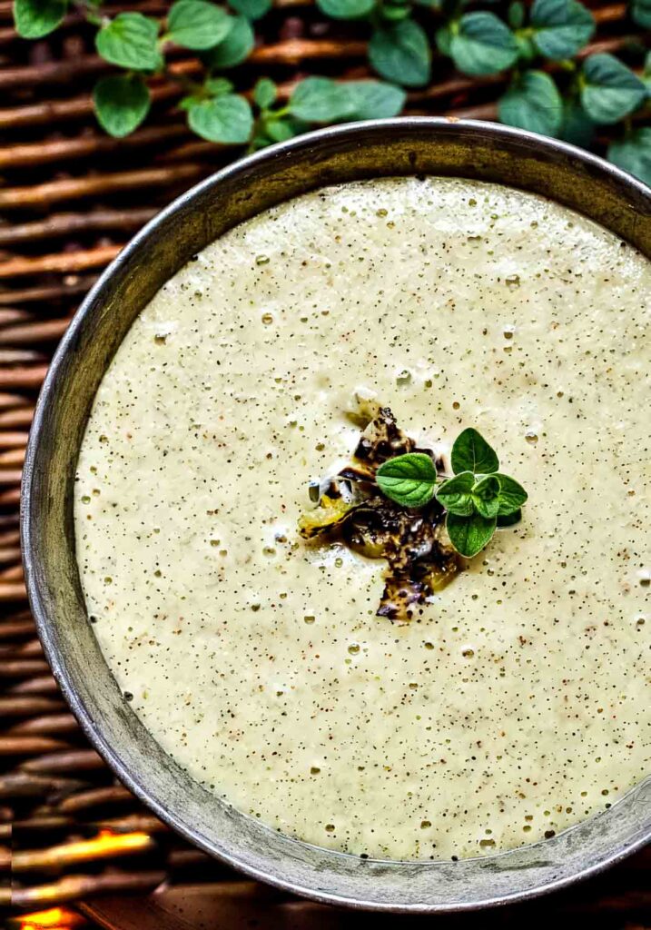 Bowl of homemade Hatch Chile Enchilada Sauce with visible chile specks, garnished with oregano and charred peppers, surrounded by fresh herbs.