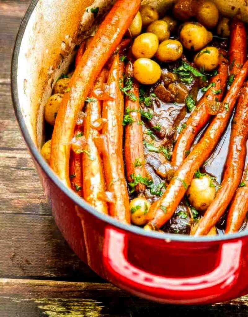 Angled shot of a red Dutch oven filled with chuck roast, carrots, and potatoes on a rustic wooden table