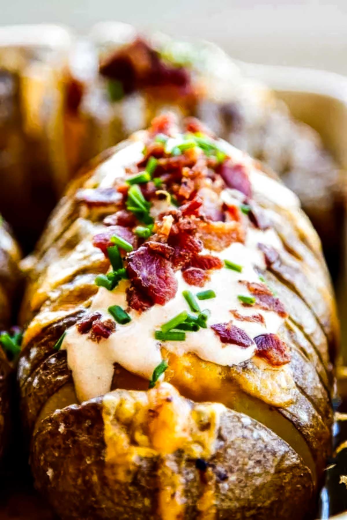a closeup of a hasselback potato topped with bacon, cheese, chipotle sour cream, and chopped chives