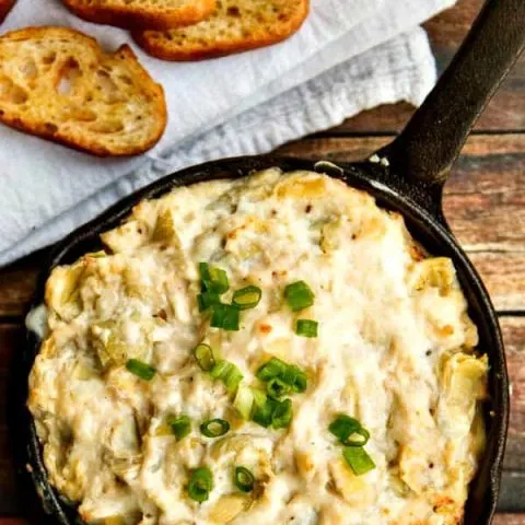 Crab and artichoke dip in a cast-iron skillet on a wood table, next to a white towel with crostini on top