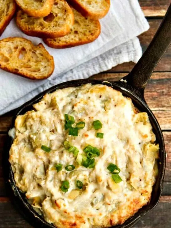 Crab and artichoke dip in a cast-iron skillet on a wood table, next to a white towel with crostini on top