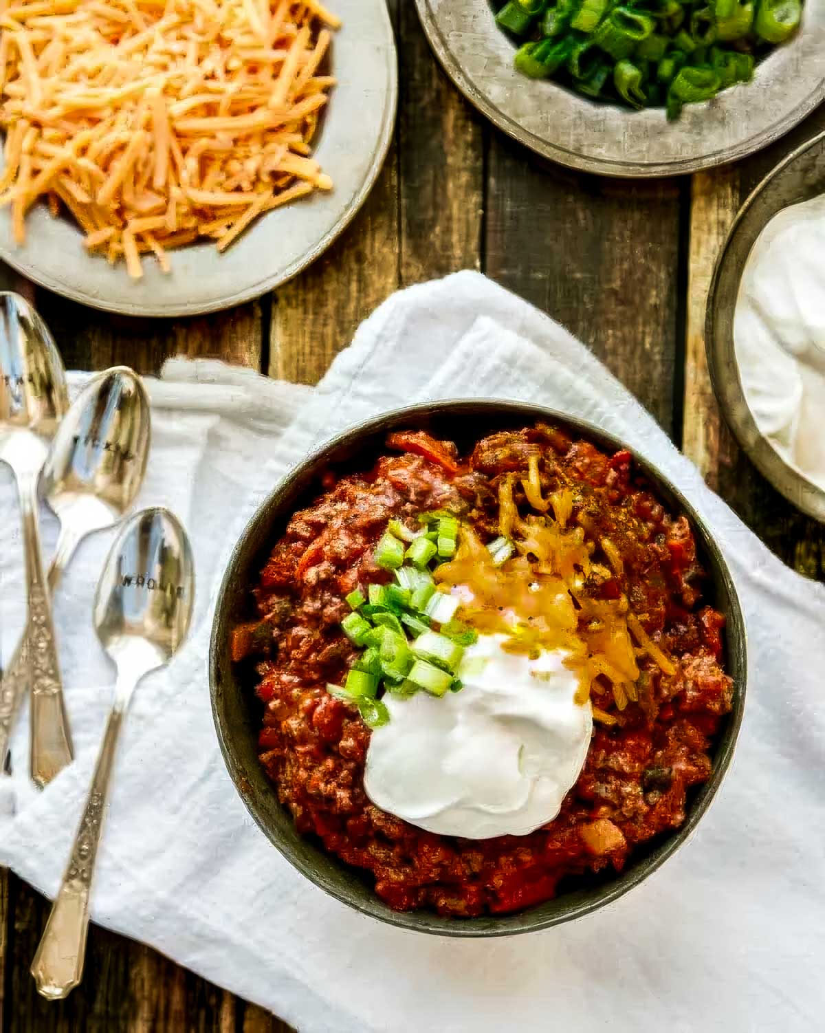 Thick and Beefy Beanless Chili Recipe a bowl of chili without beans on a white towel next to small dishes of extra shredded cheese, chopped green onion, and sour cream