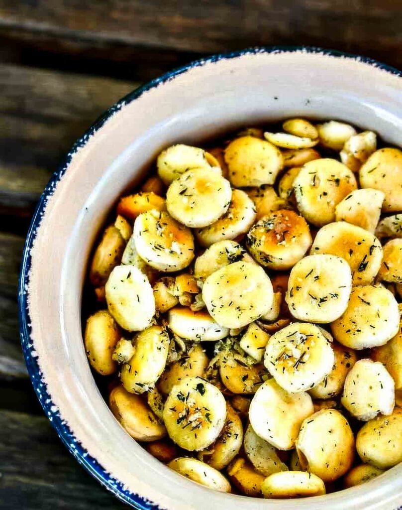 The BEST Ranch Oyster Crackers (5-Ingredient Snack)! Top-down view of seasoned ranch oyster crackers in a white ceramic bowl with blue trim, resting on a wooden table.