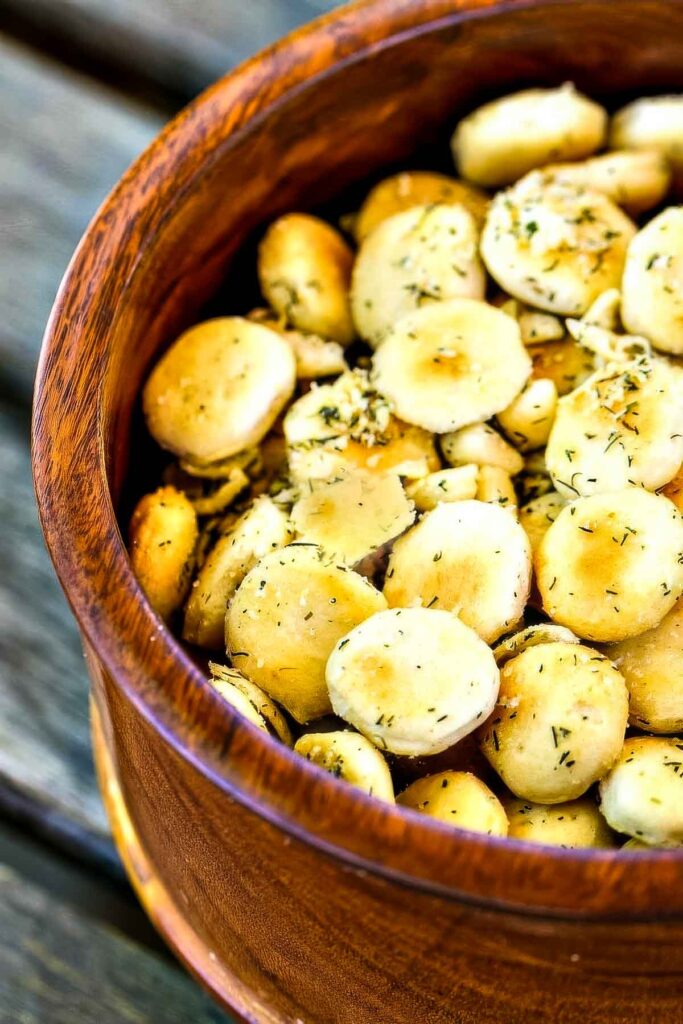Close-up of golden ranch oyster crackers in a rustic wooden bowl, topped with dried dill and garlic seasoning.