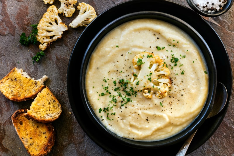 Roasted cauliflower soup in rustic black bowl, with crusty sourdough toast.