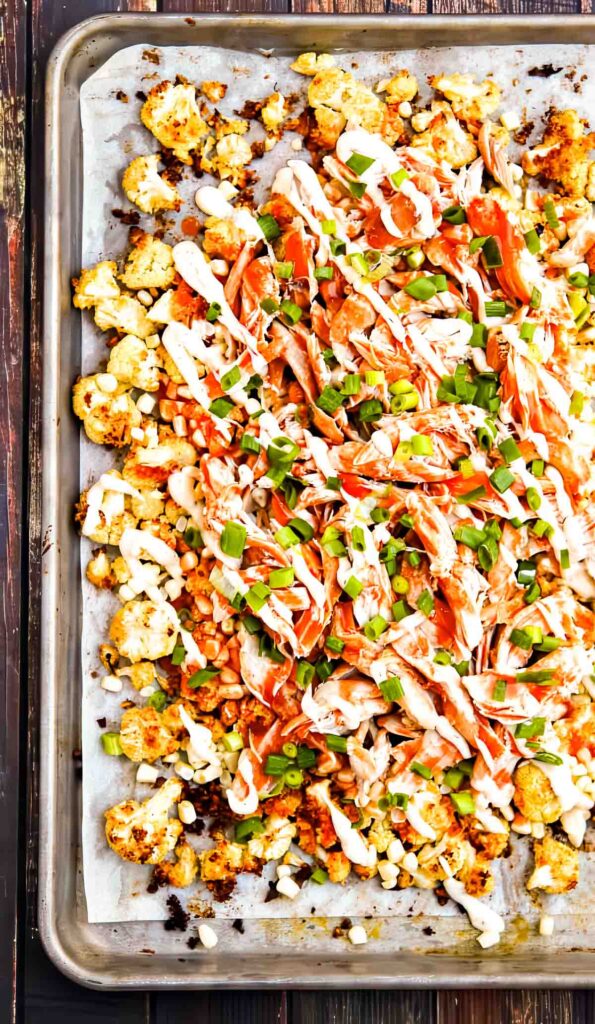 Sheet pan of roasted cauliflower nachos topped with shredded chicken, buffalo sauce, ranch dressing, and chopped green onions.