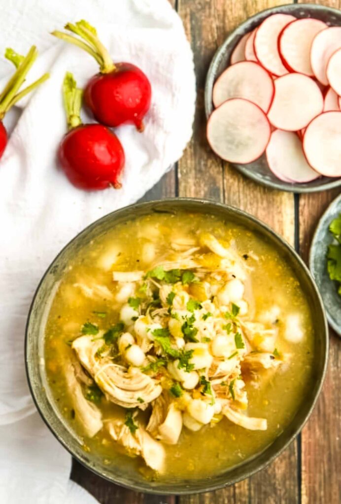 5-Ingredient Chicken Posole Recipe (Fast & Flavorful!) Top down view of a bowl of chicken posole soup with shredded chicken, hominy, and chopped cilantro in a green chile broth, surrounded by sliced radishes and whole radishes on a rustic wooden table.