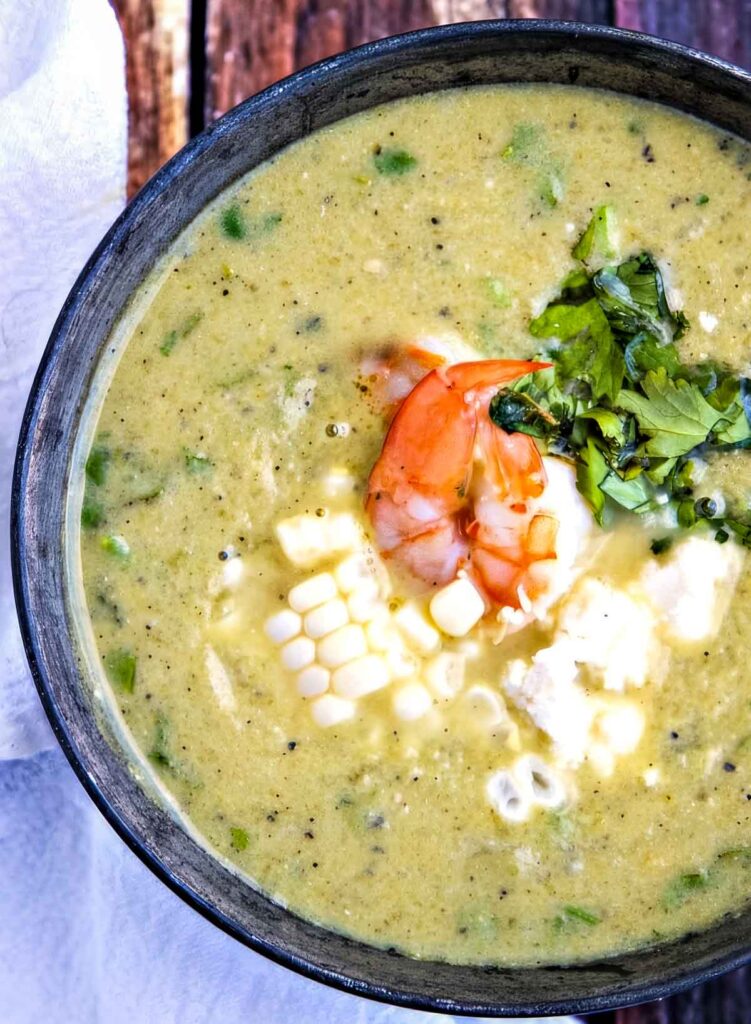 Creamy Hatch Chile Soup with Corn & Queso Fresco a closeup overhead shot of a bowl of hatch chile soup topped with shrimp and garnished with chopped parsley on a rustic wooden table
