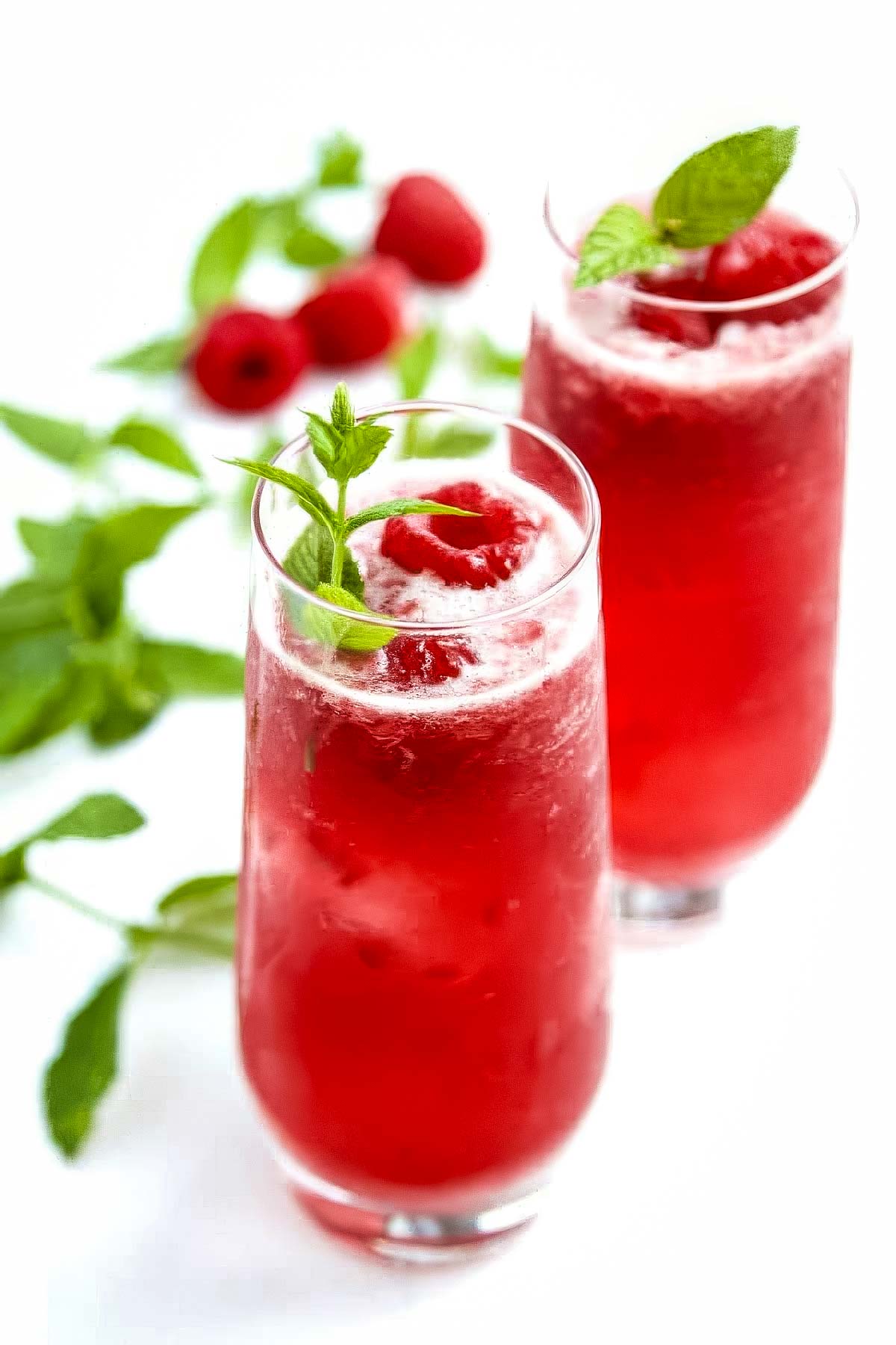 two gorgeous PAMA lemonade cocktails with a fresh raspberry and herb sprig on a white table