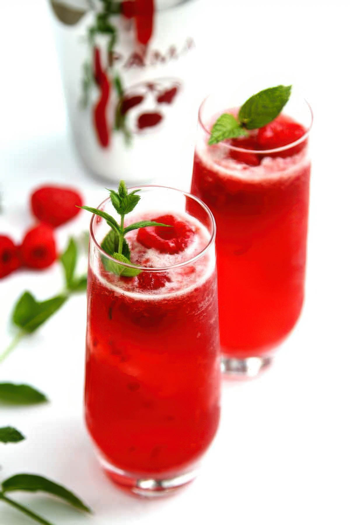 two gorgeous PAMA cocktails with a fresh raspberry and herb sprig on a white table with a bottle of PAMA liqueur in the background