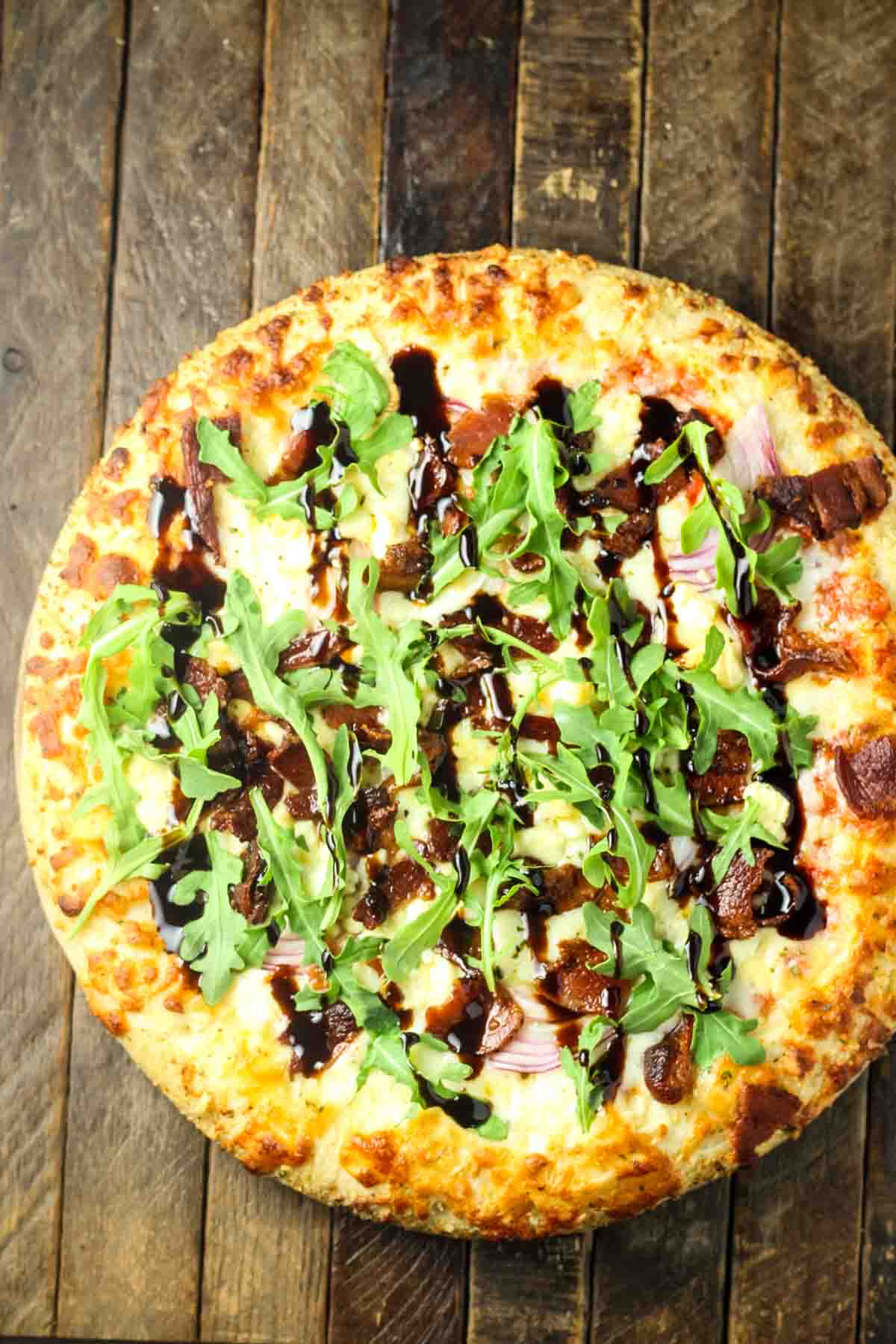 Blue Cheese Pizza Recipe – Quick Weeknight Dinner Idea an overhead shot of a frozen cheese pizza with some sliced red onion, crumbled blue cheese, crispy bacon, balsamic glaze, and fresh arugula added at home, it's just out of the oven and placed on a wooden table