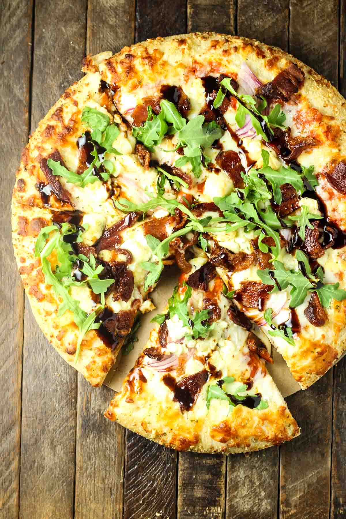 Blue Cheese Pizza Recipe – Quick Weeknight Dinner Idea an overhead shot of a frozen cheese pizza with some sliced red onion, crumbled blue cheese, crispy bacon, balsamic glaze, and fresh arugula added at home, it's just out of the oven and placed on a wooden table, it has one slice cut out