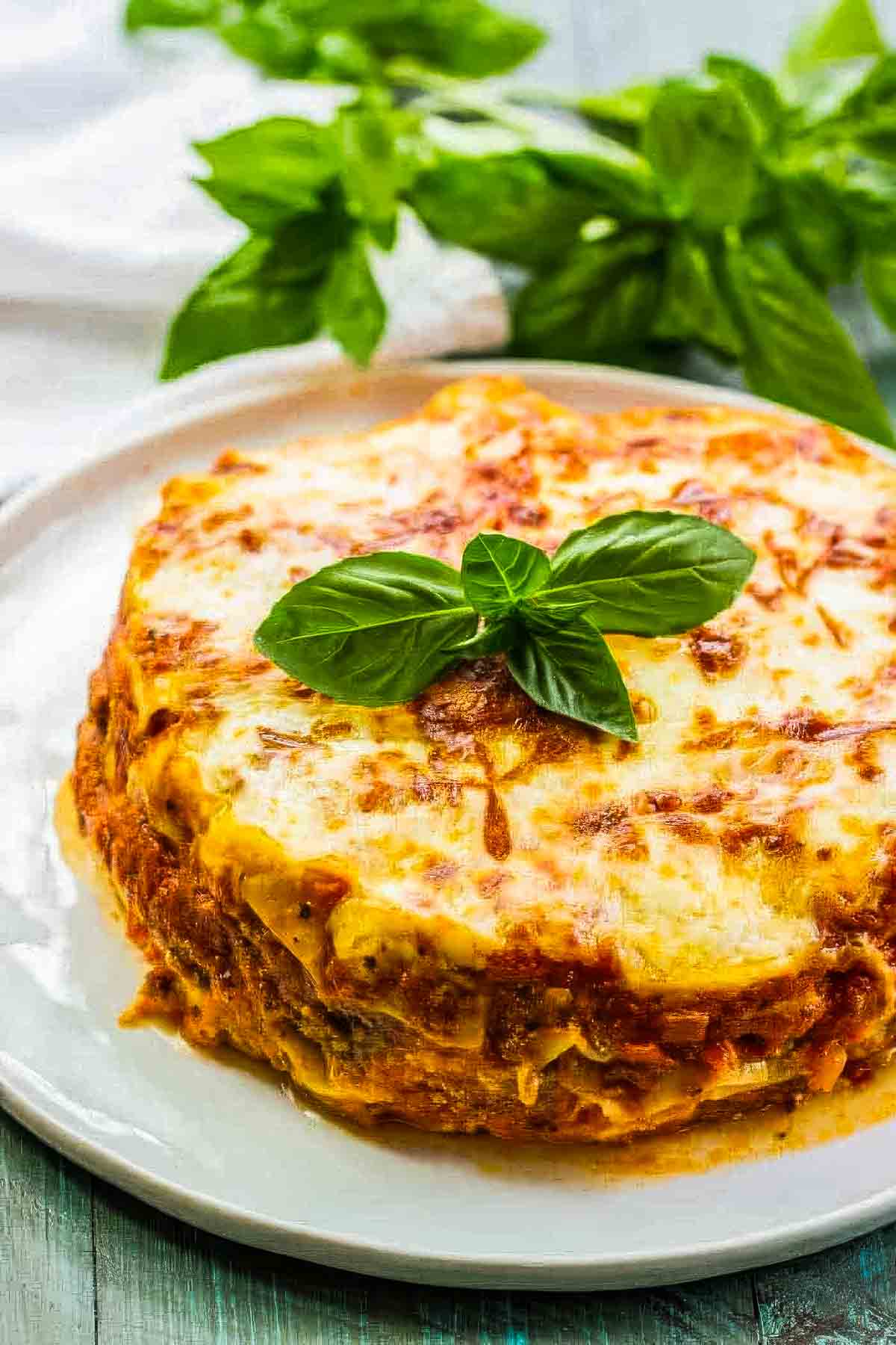 Instant Pot Lasagna (Easy, Cheesy, and Ready in 30 Minutes) instant pot lasagna on a white plate garnished with fresh basil, with more fresh basil and a white cloth in the background