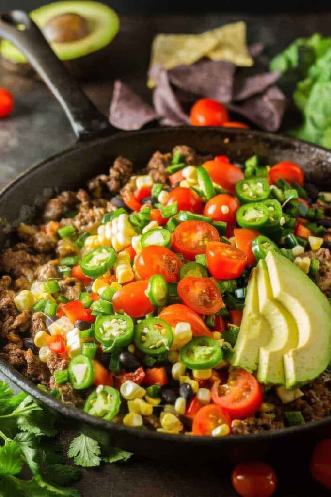 Side view of a cast iron pan with healthy nachos topped with fresh vegetables and avocado slices, surrounded by ingredients like cherry tomatoes, cilantro, and an avocado half.