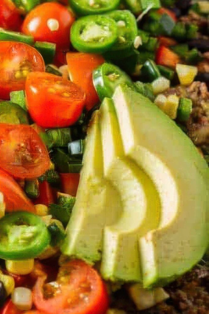 Extreme close-up of fresh avocado slices surrounded by chopped cherry tomatoes, jalapeños, green onions, and corn atop loaded healthy nachos.