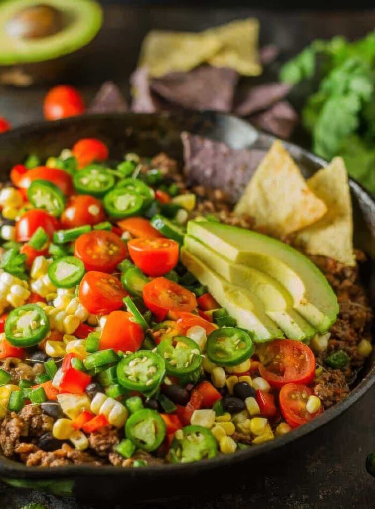 Close-up of a colorful skillet filled with healthy nachos topped with sliced avocado, cherry tomatoes, jalapeños, corn, black beans, and ground beef, served with tortilla chips.