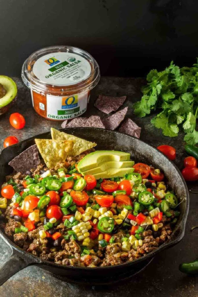 Overhead shot of a cast iron skillet filled with healthy nachos made with seasoned ground beef, fresh vegetables, and avocado, with blue corn and yellow tortilla chips on the side.