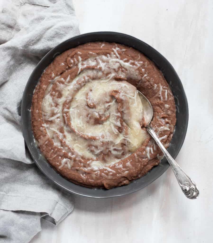 The BEST Instant Pot Refried Beans A black bowl filled with creamy Instant Pot refried beans and topped with melted white cheese, on a white tablecloth with a pretty gray napkin.
