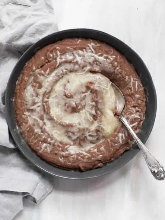 A black bowl filled with creamy Instant Pot refried beans and topped with melted white cheese, on a white tablecloth with a pretty gray napkin.