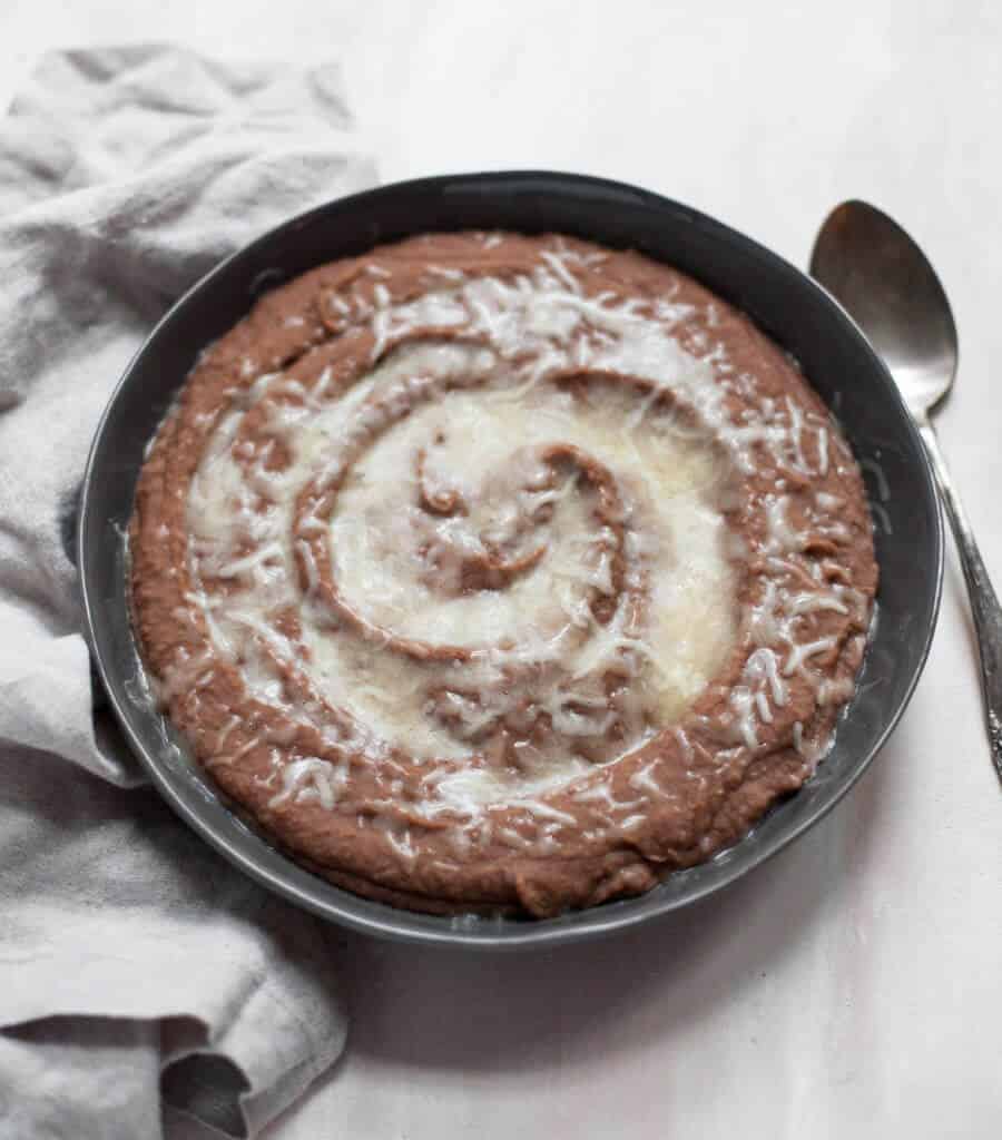 The BEST Instant Pot Refried Beans A black bowl filled with creamy Instant Pot refried beans and topped with melted white cheese, on a white tablecloth with a pretty gray napkin.