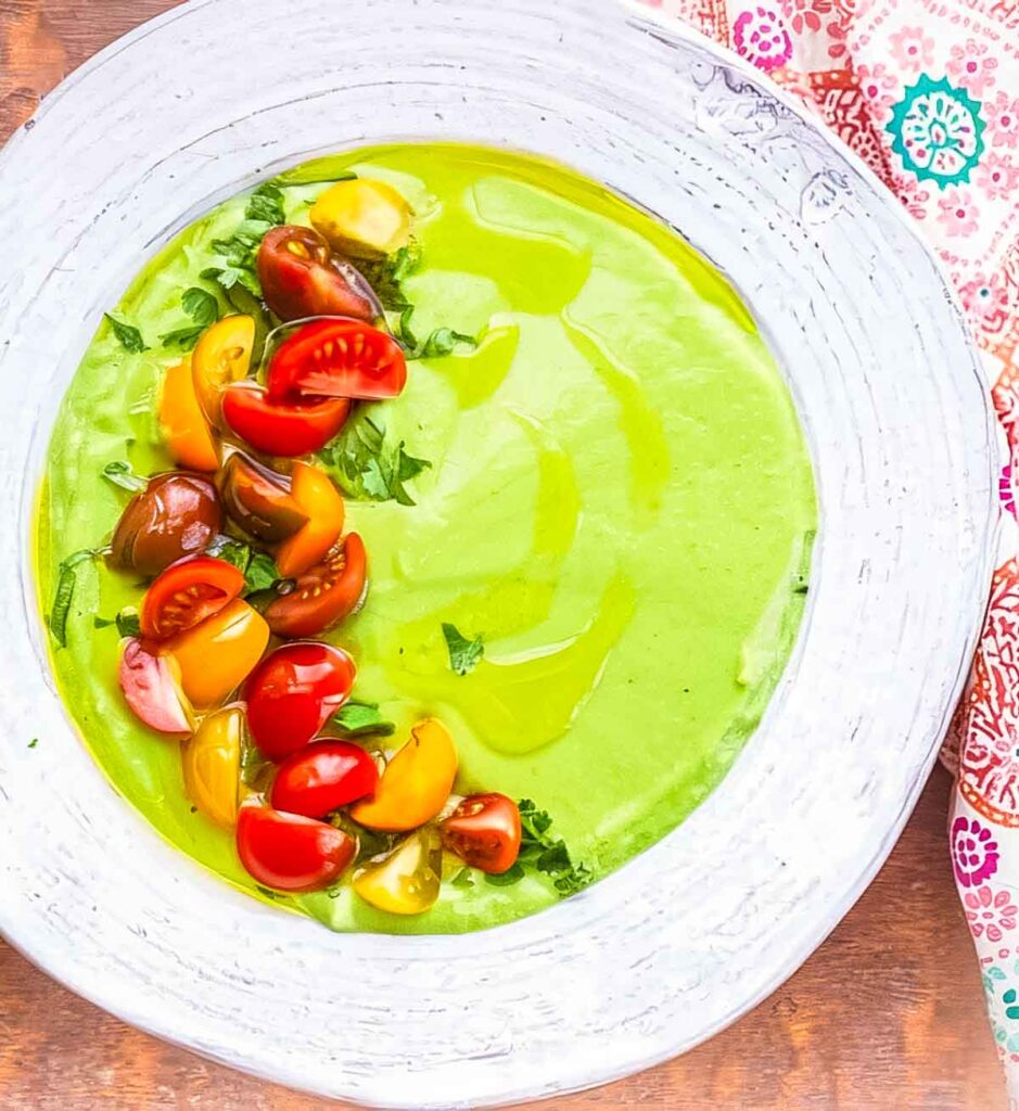 a white bowl filled with avocado soup with quartered cherry tomatoes and a drizzle of olive oil garnishing the left half of the soup
