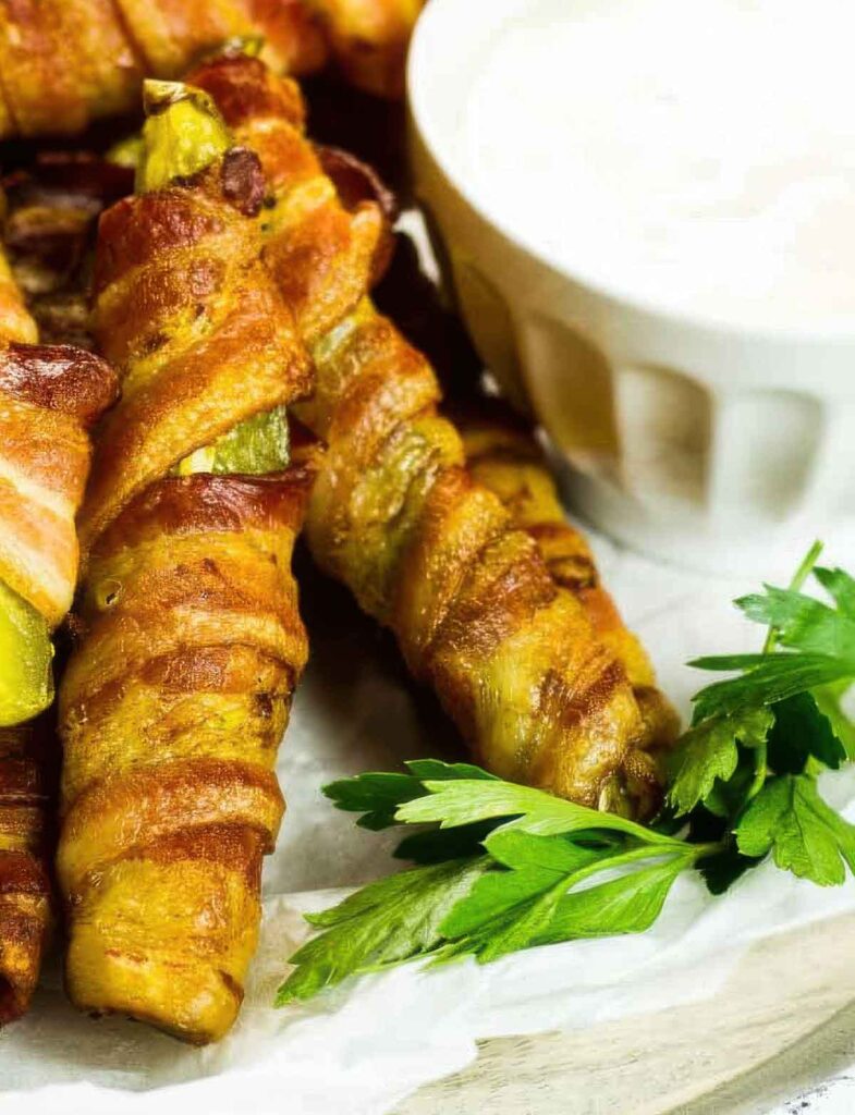 How to Make Bacon-Wrapped Pickles (Only 2 Ingredients!) Close-up of crispy bacon-wrapped pickle spears on parchment paper, served with a side of creamy dipping sauce and garnished with fresh parsley.