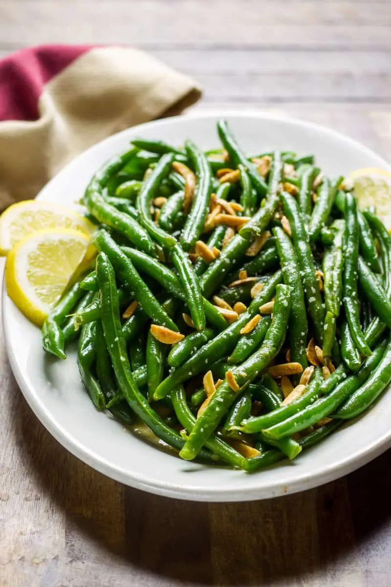 Green Beans Almondine on a white oval plate which sits on a wooden board with a rustic napkin next to it.