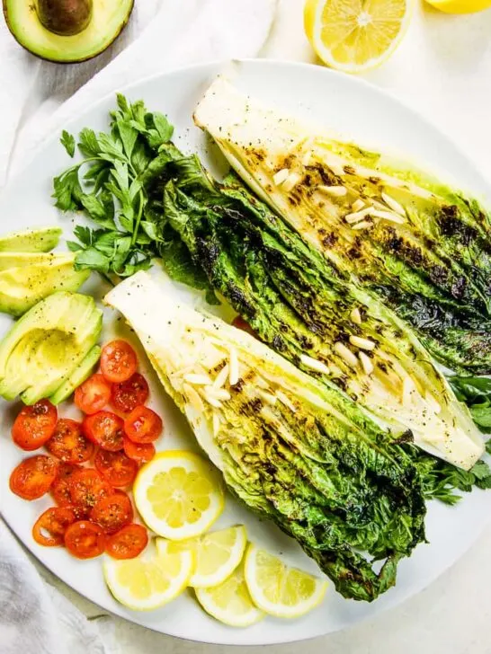 A keto salad on a white plate, made up with grilled romaine, sliced cherry tomatoes, thinly sliced avocado and halved lemon slices for a garnish.