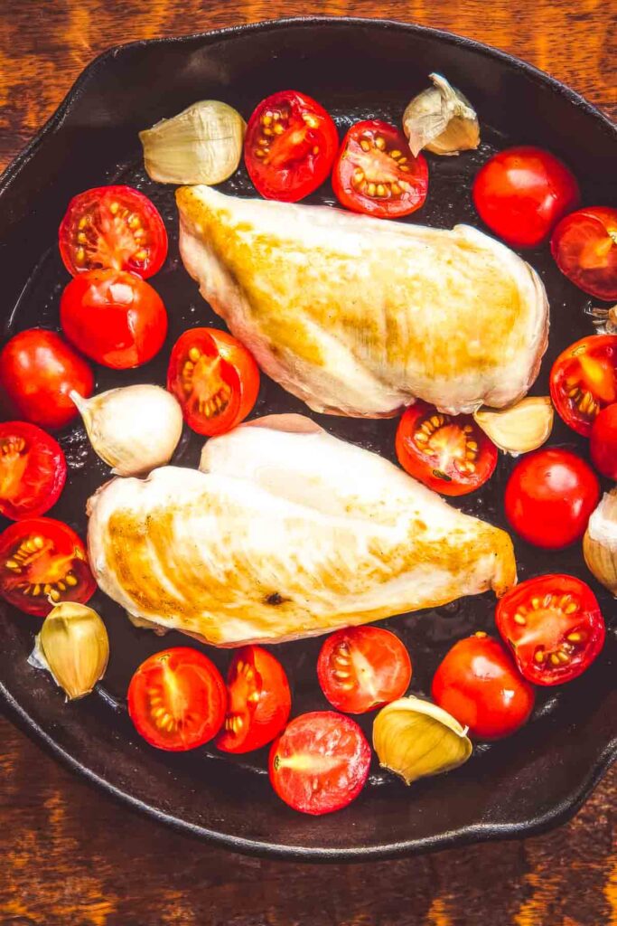 Air Chilled Chicken: Why It’s Better + A Recipe two air chilled chicken breasts being seared in a cast iron skillet with halved cherry tomatoes and whole garlic cloves