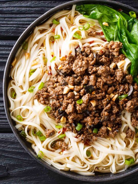 A Plate of Szechuan Gourmet Beef Pasta on a Black Table with Greens and Chopped Peanuts