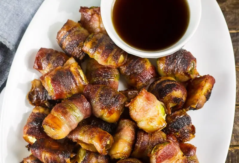 Bacon Wrapped Brussels Sprouts with Maple Syrup on a white plate next to a white bowl filled with additional maple syrup.