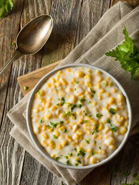 A bowl of creamed corn on a brown napkin and a wooden board