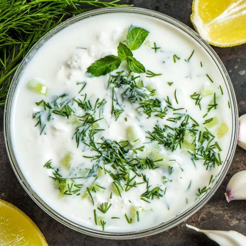 a closeup of a bowl of tzatziki sauce with fresh lemon, garlic cloves, and fresh dill and mint around the bowl on a table